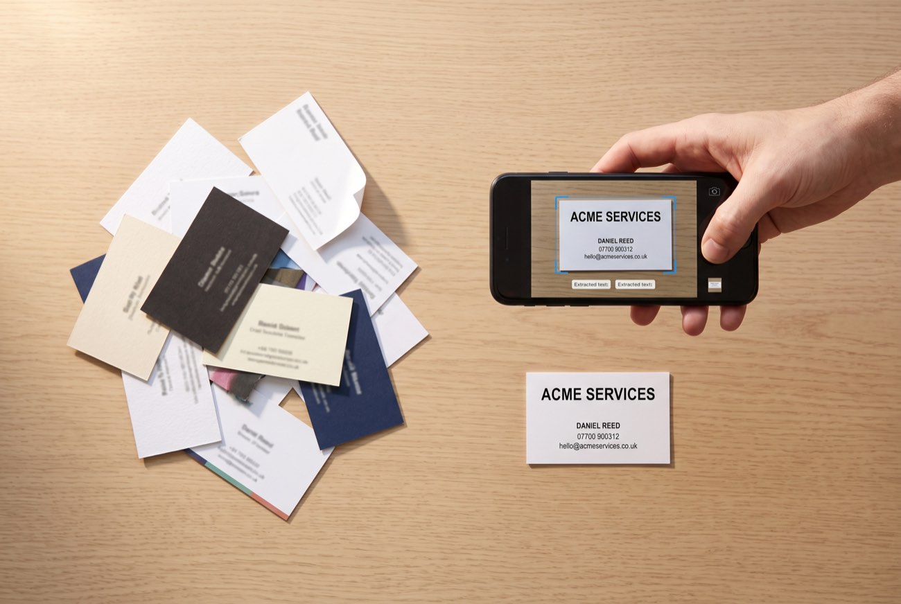 A phone held above a business card, scanning it into contacts, beside a shuffled pile of cards collected at a trade show